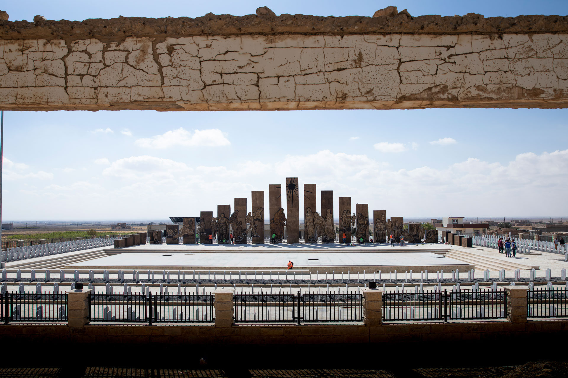 A Yazidi memorial in Sinjar: tall stone slabs carved with figures of the dead, the tallest bearing the Yazidi sun, standing over rows of white grave markers, seen from inside a building destroyed in the attack.